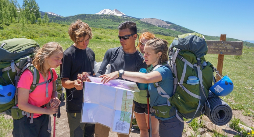 A group of people wearing backpacks examine a map. In the distance, there is a snow-capped mountain. 